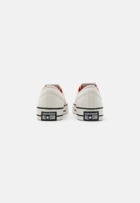 Zapatillas de lona blancas con acentos naranjas, que cuentan con una suela de goma, un bucle trasero y la marca "Converse All Star" en el talón.