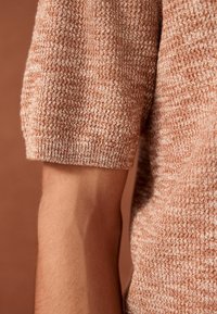 Gros plan sur un avant-bras portant un pull en maille à manches courtes texturé dans des tons marron clair et beige mélangés, sur un fond marron.