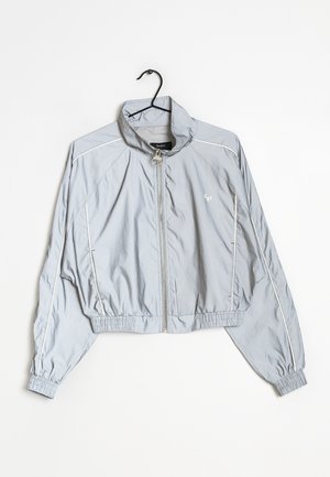 Veste coupe-vent courte gris clair en matériau lisse et léger. Présente un col haut, une fermeture éclair et des poignets élastiques.