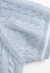 Tissu tricoté bleu clair avec un motif de point de câble, montrant la texture et les bords pliés sur un fond blanc.