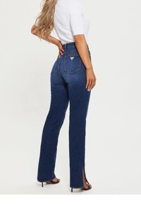 Jeans de mezclilla oscuros de cintura alta con forma de pierna recta, que presentan aberturas laterales en el dobladillo y un logo triangular en el bolsillo trasero.