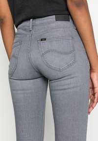 Jeans de mezclilla gris con un diseño clásico, bolsillos traseros y una etiqueta de parche de cuero negro en la cintura. Textura suave y forma ajustada.