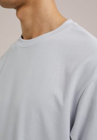 Svetlo siva majica s okruglim izrezom, napravljena od mekog materijala, s opuštenim krojem i glatkom teksturom, koja prikazuje minimalne šavove i nema vidljivih uzoraka.