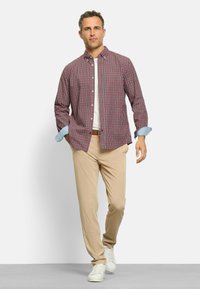 Mann, der ein rot-kariertes Hemd über einem weißen T-Shirt, beige Hosen, braemen Gürtel und weiße Sneaker trägt, vor einfarbigem Hintergrund.