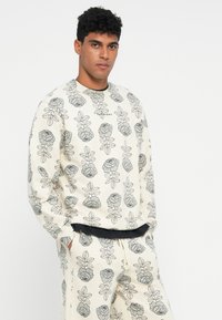 Hombre con sudadera color crema y pantalones con un patrón floral gris, de pie con las manos en los bolsillos contra un fondo blanco.