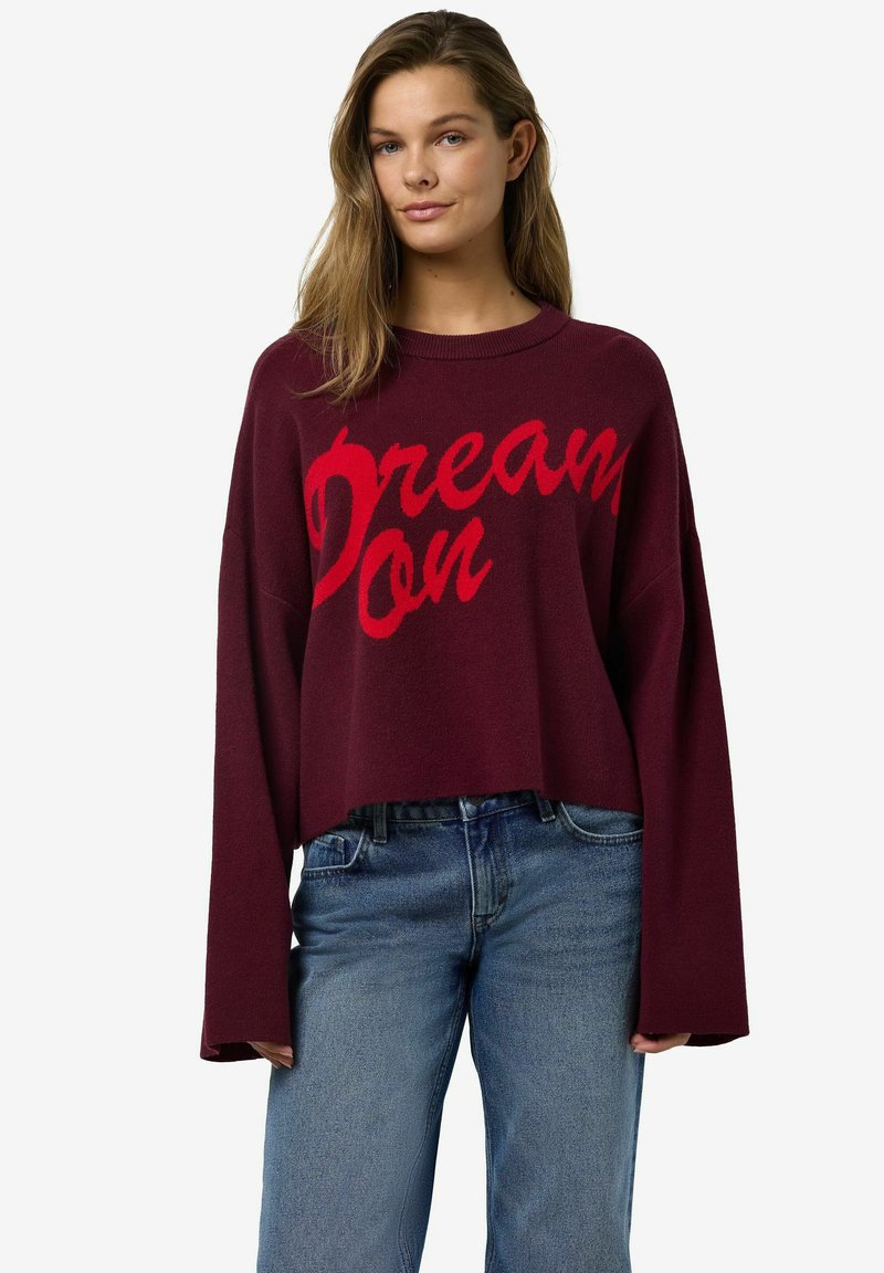 Burgundy cropped sweater with a loose fit, featuring a red "Dream On" text design. Soft knit texture and wide, long sleeves. Paired with blue jeans.