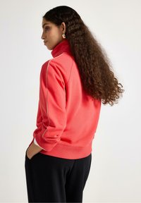 Felpa color corallo con collo alto, caratterizzata da piping bianco a contrasto lungo le spalle e polsini a costine. Tessuto morbido e vestibilità morbida.
