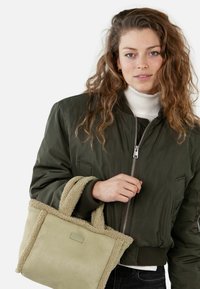 Olivgrüne Bomberjacke mit silbernem Reißverschluss, getragen über einem weißen Rollkragenpullover. Das Modell hält eine hellgrüne, strukturierte Tragetasche mit weichem, flauschigem Besatz.