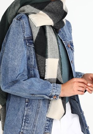 Personne portant une veste en jean bleue, un haut vert, un pantalon blanc et une grande écharpe à carreaux noire, grise et crème autour du cou.