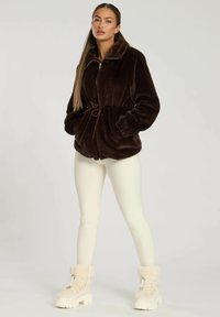 Bruine nep-bontjas met een hoge kraag, een trekkoord in de taille en zijzakken, gecombineerd met witte leggings en crèmekleurige laarzen met bontvoering.