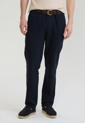 Mężczyzna noszący granatowe spodnie o prostych nogawkach, brązowy pasek ze złotą klamrą, czarne wsuwane buty oraz kremowy krótkim sweter w warkoczowy splot.