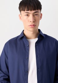 Cămașă bleu închis, cu nasturi, confecționată dintr-un material neted, având guler și închidere cu nasturi la față, suprapusă peste un tricou alb.