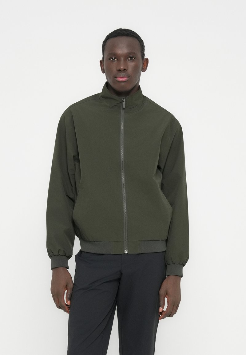 Νέος άνδρας που στέκεται φορώντας ένα σκούρο πράσινο μπουφάν με φερμουάρ και μαύρο παντελόνι πάνω σε ένα απλό λευκό φόντο.