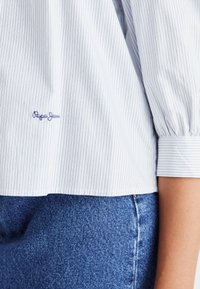 Balti ir mėlyni dryžuoti marškiniai su pūstomis rankovėmis, su išsiuvinėtu "Pepe Jeans" logotipu. Suderinti su mėlynais džinsais.