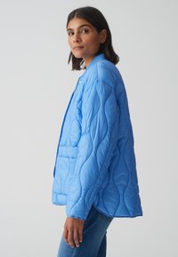 Veste bleue matelassée avec un col structuré, des poches latérales et un motif de couture visible. Le tissu semble lisse et léger.