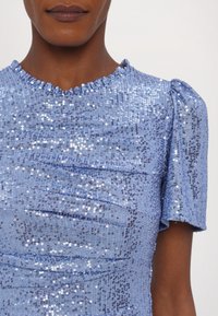 Femme portant un haut bleu clair à manches courtes recouvert de petites paillettes brillantes, avec un col volanté et des plis horizontaux texturés.