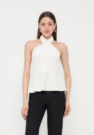 Donna che indossa un top bianco smanicato con collo halter e orlo a peplo e pantaloni neri su uno sfondo chiaro e semplice.