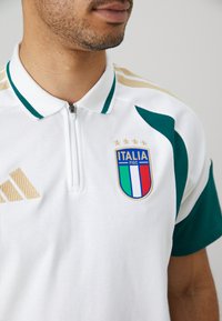 Hvit polo-shirt med grønne detaljer, har en glidelås krage. Inkluderer et brodert Italia-merke med tre stjerner og gull Adidas-logo.