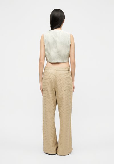 Γυναίκα με μακριά σκούρα μαλλιά που φοράει αμάνικο ριγέ crop top και φαρδιά μπεζ παντελόνια με μικρές τρύπες, στεκόμενη με την πλάτη προς τα εμάς.