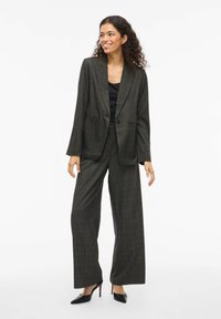 Tamsiai žalias languotas kostiumas su rudos spalvos akcentais. Švarkas turi vieno mygtuko užsegimą, įpjautas apykakles ir laisvą siluetą; pločio kojų kelnės užbaigia įvaizdį.