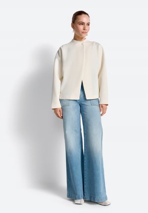 Naine seisab käed külgedel, seljas kreemikas pikkade varrukatega top, helesinised laiade säärega teksapüksid ja valged teravnurksed kingad.