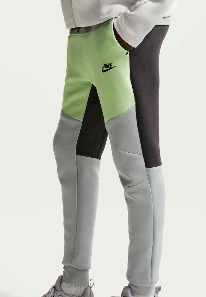 Person iført farveblok Nike joggingbukser i grøn, sort og grå, stående med hænderne i lommerne og synlige grå sneakers.