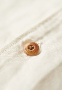 Tessuto beige chiaro con un grande bottone marrone impresso con la scritta "SUPERDRY". Il bottone presenta un foro centrale e una texture rigata.