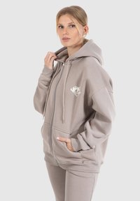 Grijze zip-up hoodie met een grote kangoeroezak, ribgebreide boorden en een klein logo op de borst. Zacht materiaal en een relaxte pasvorm.