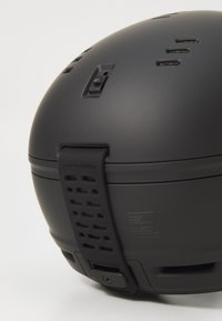 Svart hjelm med matt finish, med ventilasjonsåpninger, justerbar stropp og preget merkevare. Glatt overflate med rund form.