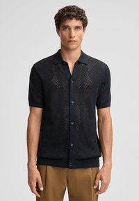 Polo shirt nero a maniche corte con un motivo testurizzato a trama aperta. Presenta un colletto tradizionale e una chiusura con bottoni. Completato da un orlo scuro.