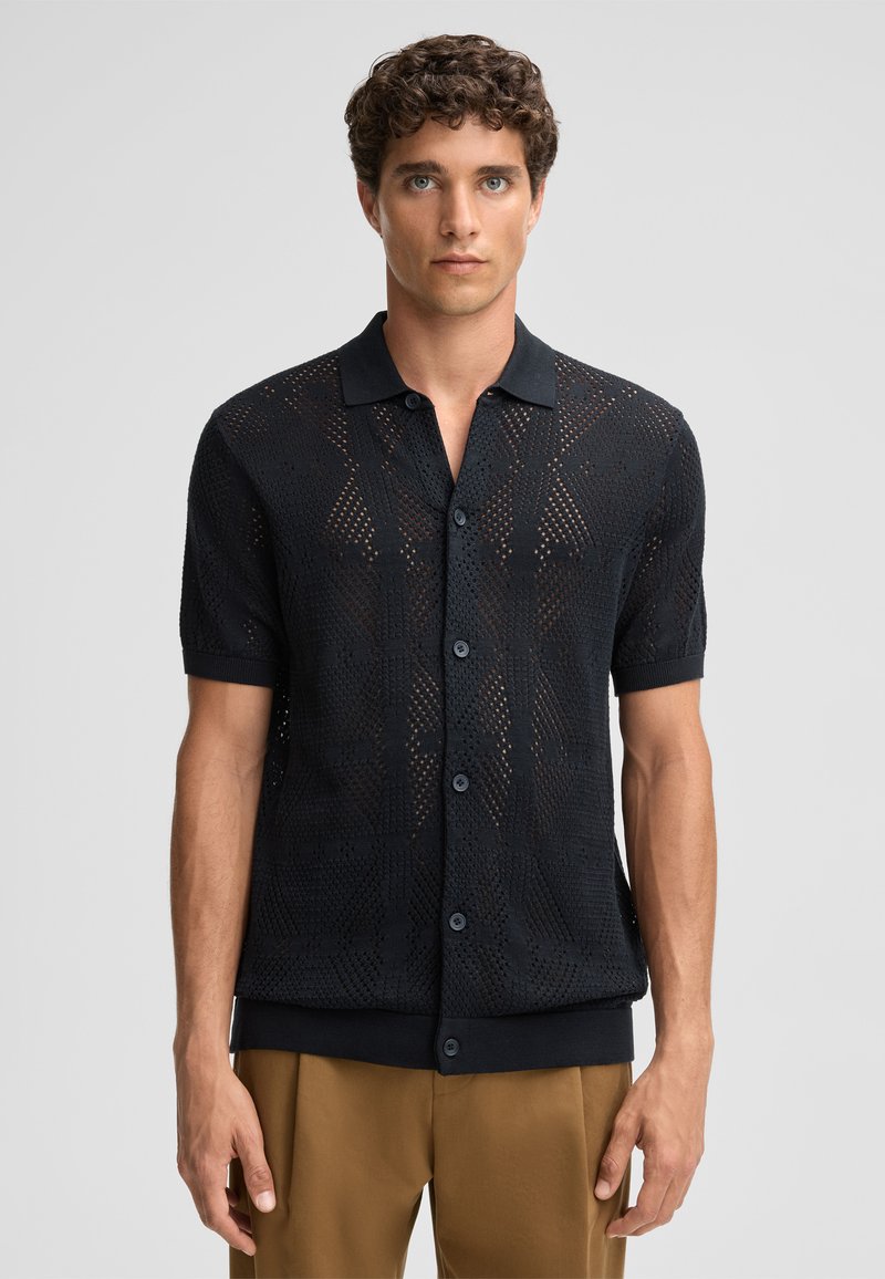Polo shirt nero a maniche corte con un motivo testurizzato a trama aperta. Presenta un colletto tradizionale e una chiusura con bottoni. Completato da un orlo scuro.