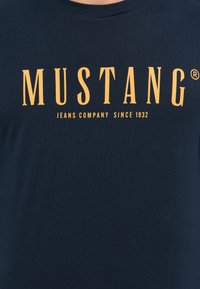 Tengerészkék pamut póló, sárga felirattal, amely így szól: "MUSTANG JEANS COMPANY SINCE 1932", modern serif betűtípussal.