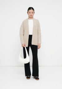 Ung kvinna i beige stickad cardigan, vit topp, svarta utsvängda byxor, svarta skor, håller en vit väska, står mot vit bakgrund.