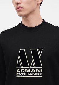 Νέος άνδρας που φοράει μαύρο φούτερ Armani Exchange με μεγάλο λευκό και μαύρο λογότυπο στο στήθος, στραμμένος ελαφρώς αριστερά.