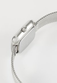 Montre en métal argenté avec un boîtier rectangulaire, une couronne texturée et un bracelet en maille. L'arrière présente des détails de marque gravés.