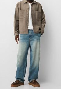 Fleece-Hemd mit Knöpfen in Taupe, zwei Brusttaschen. Hellblaue Jeans mit weitem Bein und verwaschenem Wascheffekt. Braune Schuhe mit dicken Sohlen.
