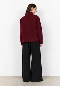 Bordeaux fleece jack met een hoge kraag, lange mouwen en een relaxed fit, gecombineerd met wijde zwarte broek. De stof lijkt zacht en gestructureerd.