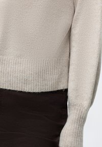 Pull beige en tricot avec poignets et ourlet côtelés porté sur un pantalon noir, montrant le côté et le bas du torse.