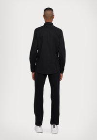 Camicia nera con bottoni, maniche lunghe, texture liscia e taglio dritto, abbinata a pantaloni neri e sneakers bianche.
