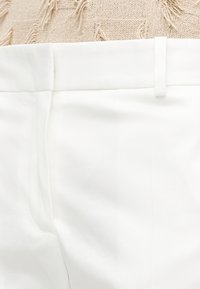 Κοντινή λήψη της μέσης λευκών παντελονιών με θηλιά ζώνης πάνω σε υφή μπεζ ύφασμα.