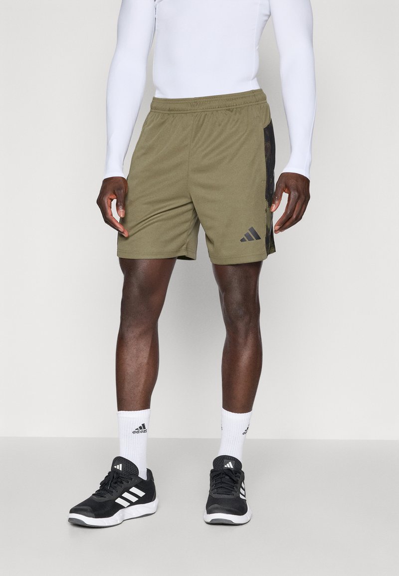 Žalios alyvuogių spalvos šortai su elastinga juosta, turintys juodą šoninį panelį ir Adidas logotipą. Suderinti su juodais sportiniais batais ir baltais kojinaitėmis.