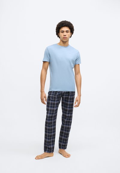 Tricou bleu deschis cu mâneci scurte, asortat cu pantaloni scurți în carouri în nuanțe de albastru, negru și alb. Modelul stă desculț pe un fundal neutru.