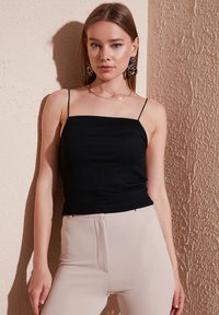 Čierny rebrovaný top na tenkých ramienkach, nosený s béžovými priliehavými nohavicami. Pozadie so štruktúrovanou béžovou stenou. Celkový elegantný dizajn.