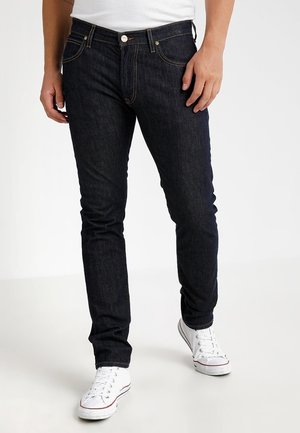 Homme portant un jean slim foncé et des baskets blanches, debout les mains relâchées le long du corps.