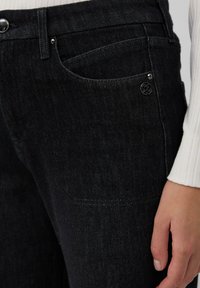 Des jeans en denim noir présentent un design ajusté avec des poches visibles, un bouton argenté et des détails de couture subtils. La texture semble lisse et durable.