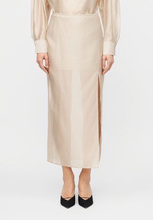 Personne portant une jupe midi beige transparente avec une fente latérale, associée à un haut à manches longues et des escarpins noirs à bouts pointus.