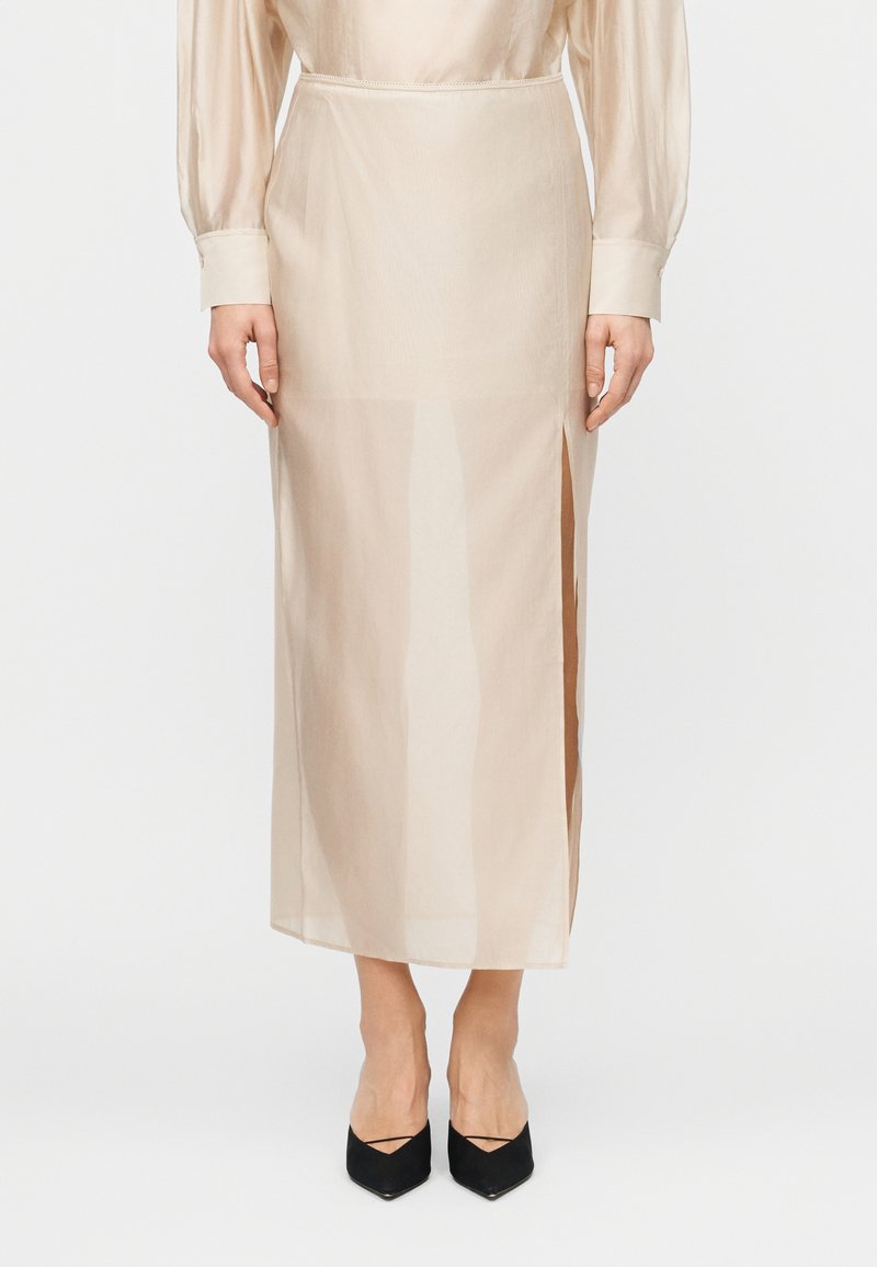 Persoană purtând o fustă midi bej transparentă cu o deschizătură laterală, combinată cu un top cu mâneci lungi și pantofi negri cu vârf ascuțit.