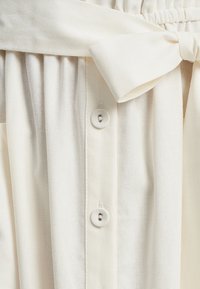 Light cream-colored garment with a belted waist, featuring pleats and button closures. Made from a soft, textured fabric.