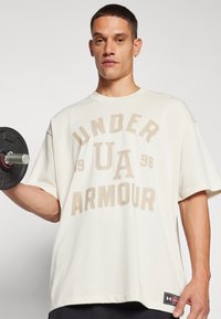 Camiseta blanca de gran tamaño con el logo de "Under Armour" en beige. El modelo sostiene un peso negro. Manga corta, escote redondo y etiqueta de marca en el dobladillo.