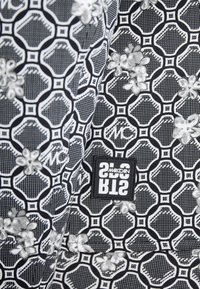 Schwarz-weiß gemustertes Gewebe mit floralen Designs und geometrischen Formen. Enthält ein schwarzes Etikett mit weißem Text.